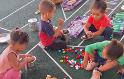 #paratodosverem: Quatro crianças estão sentadas no chão de um pátio, que tem marcações esportivas, brincando com materiais reciclados. Elas interagem com latas de metal, garrafas plásticas, papelão, pedras e tampinhas coloridas espalhadas pelo chão, em uma atividade lúdica que incentiva a criatividade e o uso de materiais não estruturados