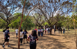 #paratodosverem: Um grupo de jovens, a maioria de costas, caminha em uma trilha de terra sob a copa de árvores secas e frondosas, em um parque. A cena, vista por trás de um tronco, mostra os participantes de mochilas, em um momento de lazer e aprendizado ao ar livre.