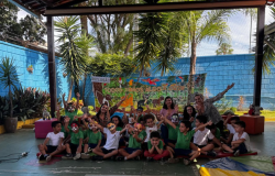 #paratodosverem: Crianças e educadoras posam sorrindo em um pátio aberto, muitas delas usando pintura facial de animais. Atrás, um painel colorido com desenhos da fauna e a frase “Todos temos algo a oferecer”. 