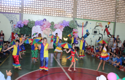 #paratodosverem: Na foto, crianças se apresentam em um ginásio escolar decorado com balões coloridos. Elas usam fantasias vibrantes em azul, amarelo, vermelho e verde, e seguram sombrinhas multicoloridas, típicas de danças populares brasileiras. Com sorrisos e movimentos coreografados, enchem o espaço de cor e alegria, enquanto famílias assistem animadas ao espetáculo.