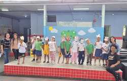 Legenda: Aprendizado e diversão no palco: arte e teatro em cena #paratodosverem: A imagem mostra um grupo de crianças em um palco decorado com um painel azul, nuvens, sol e árvores de papel. As crianças usam máscaras de porquinhos e roupas coloridas. À frente do palco, algumas crianças estão sentadas no chão sobre um tapete colorido com números. Quatro professoras sorriem junto ao grupo.