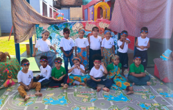 #paratodosverem: Na imagem, um grupo de crianças posa sorridente em um espaço decorado com tecidos coloridos, representando um cenário cultural. Elas usam faixas na cabeça e roupas estampadas, sugerindo uma atividade escolar temática, ligada à cultura afro-brasileira e indígena
