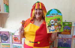 #paratodosverem: Na foto, uma mulher sorri, vestida como a personagem Emília, do Sítio do Picapau Amarelo. Ela usa uma fantasia colorida em vermelho e amarelo, com uma peruca de tiras coloridas. Em uma mão segura um livrinho do Sítio e, na outra, uma pequena boneca. Ao fundo, há prateleiras cheias de livros infantis.