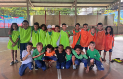 #paratodosverem: Em uma quadra coberta, um grupo grande de crianças, vestindo coletes verde-limão e laranja, está posando para uma foto em duas fileiras. Eles estão abraçados, com sorrisos e gestos de "joinha". 