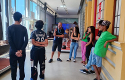 #paratodosverem: Em um corredor de museu, iluminado por grandes janelas laterais, um grupo de jovens está reunido em torno de um professor ou monitor. O monitor, de camiseta preta e crachá, gesticula enquanto conversa com o grupo. A cena, com pisos claros e paredes amarelas, transmite um momento de aprendizado e visita cultural.