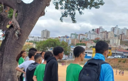 #paratodosverem: Um grupo de jovens, em sua maioria meninos, está de pé em uma área de terra, olhando para um campo de futebol abaixo e para uma paisagem urbana com muitas casas e prédios em um morro ao longe. A cena é em um dia nublado. Em primeiro plano, um tronco de árvore retorcido enquadra a vista da comunidade