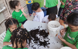 #paratodosverem: Crianças pequenas participam de uma atividade prática em grupo, explorando o conteúdo de baldes com terra e minhocas sobre uma mesa. Elas usam camisetas verdes com o logotipo da Prefeitura de Belo Horizonte.