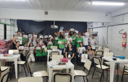 #paratodosverem: Um grupo de estudantes posa em uma sala de aula segurando trabalhos feitos com folhas e elementos naturais. Eles sorriem e mostram suas produções. Algumas professoras estão sentadas na frente, também segurando os trabalhos. Mesas e cadeiras estão espalhadas pelo espaço.