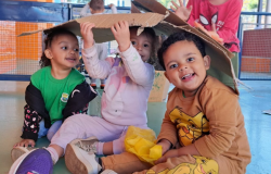 #paratodosverem: Três crianças pequenas estão sentadas no chão de um pátio, rindo e brincando sob um pedaço de papelão que usam como cabana. A menina da esquerda veste uma camiseta verde, a do meio veste rosa claro, e o menino da direita veste uma blusa laranja com desenho do Rei Leão.