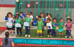 #paratodosverem: Um grupo de 15 crianças, a maioria em idade pré-escolar e vestindo uniformes verdes, está em um palco. Três crianças seguram cartazes com frases sobre a natureza, como "VAMOS CUIDAR DA NATUREZA". Ao fundo, o cenário é decorado com flores de papel. Em primeiro plano, uma professora está sentada no chão, de costas, em frente a uma faixa azul com texto.