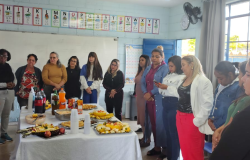 #paratodosverem: Em uma sala de aula decorada com o alfabeto, um grupo de professoras e funcionárias está reunido ao redor de uma mesa com salgados, frutas e refrigerantes. A maioria das mulheres está de pé, conversando e atenta. 
