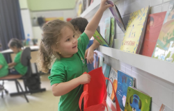 #paratodosverem: A imagem mostra uma criança pequena sorridente, usando camiseta verde, escolhendo um livro em uma estante cheia de títulos infantis. Ela segura uma sacola vermelha aberta, como se fosse guardar o livro dentro dela. Ao fundo, outras crianças também estão ocupadas na sala de aula.