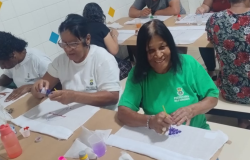 #paratodosverem: Estudantes da Educação de Jovens e Adultos participam de uma oficina de pintura em tecido. Sentados em mesas coletivas, eles utilizam tintas coloridas e pincéis para criar desenhos.