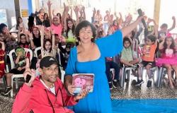 #paratodosverem: Em um espaço interno de uma escola, um homem em cadeira de rodas segurando um livro e uma mulher vestindo um vestido azul com um microfone sorriem enquanto estão à frente de um grupo de crianças sentadas em cadeiras de plástico. Todos estão levantando as mãos. Algumas crianças usam fantasias coloridas.