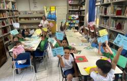 #paratodosverem: Crianças sentadas, em biblioteca escolar, mostram certificados de diferentes cores enquanto sorriem ao redor de mesas com livros e atividades. Ao fundo, há estantes com livros e uma educadora acompanha o grupo.