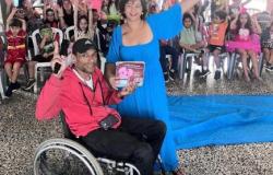 #paratodosverem: Em um espaço interno de uma escola, um homem em cadeira de rodas segurando um livro e uma mulher vestindo um vestido azul com um microfone sorriem enquanto estão à frente de um grupo de crianças sentadas em cadeiras de plástico. Todos estão levantando as mãos. Algumas crianças usam fantasias coloridas.