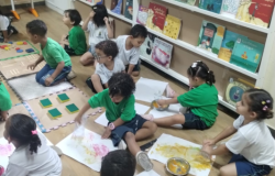 #paratodosverem: Crianças pequenas sentadas no chão de madeira de uma sala de aula participam de uma atividade artística. Elas usam potes com tinta amarela e rosa para pintar em grandes folhas de papel espalhadas pelo chão. Algumas usam uniforme verde e outras, branco. Ao fundo, uma estante branca exibe diversos livros infantis virados para a frente, e há tapetes sensoriais dispostos à esquerda.