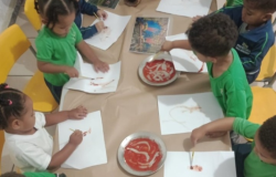 #paratodosverem: crianças pequenas sentadas ao redor de uma mesa longa com cadeiras amarelas pintam em folhas brancas. Elas usam pincéis e tinta avermelhada (urucum) disposta em dois pratos de metal sobre papel pardo. Existem objetos de madeira e fotos na mesa. Várias vestem camisetas verdes de uniforme e uma usa jaqueta azul marinho com logotipo. O chão é de lajotas.