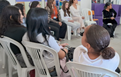 #paratodosverem: Diversas pessoas, em sua maioria jovens, estão sentadas em cadeiras brancas de plástico dispostas em círculo durante uma reunião. Em primeiro plano, três participantes aparecem de costas ou de perfil. Ao fundo, outras mulheres escutam atentas, incluindo uma com casaco em estampa de oncinha e outra vestida de preto em um sofá roxo. O ambiente tem paredes claras e cartazes coloridos.
