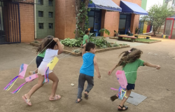 #paratodosverem: Cinco crianças correm em um pátio escolar de terra, brincando com rabiolas coloridas presas a cordões que seguram pelas mãos. Três delas correm para a direita, com fitas rosa, amarela e coloridas voando atrás. Ao fundo, há um edifício com tijolos à vista e toldos azuis sobre portas e janelas. Canteiros com plantas verdes e um banco de madeira também estão visíveis.