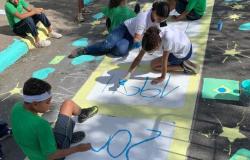 #paratodosverem: Estudantes participam da pintura de uma rua decorada para a Copa do Mundo. Sentados no chão, eles utilizam tinta azul para escrever em espaços demarcados”. Ao redor, aparecem desenhos de estrelas, bandeiras do Brasil e círculos em verde, amarelo e azul.