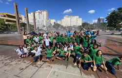 na imagem, um grupo grande de crianças e pessoas adultas está reunido em um espaço público ao ar livre, com roupas molhadas e expressões de alegria.