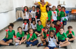 #paratodosverem: Diversas crianças de uniforme escolar posam sentadas e em pé em uma sala de aula iluminada. No centro, uma mulher vestida como a princesa Bela, de vestido amarelo, faz uma pose divertida. A maioria das crianças segura pequenos corações de papel vermelho em direção à câmera, sorrindo. Ao fundo, há um quadro branco, uma bancada de granito com pia e cadeiras organizadas na sala.