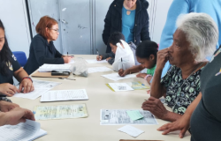 #paratodosverem: Adultos e crianças ao redor de uma grande mesa branca preenchem formulários. Em destaque à direita, uma idosa negra de cabelos grisalhos observa pensativa. No centro, duas crianças escrevem concentradas sob a supervisão de uma mulher. À esquerda, uma funcionária ruiva de óculos manuseia papéis. Ao fundo, há armários metálicos cinzas em uma sala de atendimento.