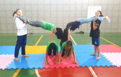 #paratodosverem: Em uma quadra esportiva com piso nas cores verde e vermelho, seis jovens realizam uma formação acrobática. No centro, duas estudantes estão de joelhos e mãos apoiadas sobre tatames coloridos de encaixe (nas cores azul, rosa e amarelo). Sobre as costas delas, outras duas fazem paradas de mão, tendo as pernas seguradas horizontalmente por duas jovens em pé nas laterais. Todas vestem uniformes.