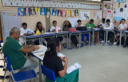 #paratodosverem: Em uma sala com paredes de tijolos e piso cinza, adultos e jovens estão sentados em mesas azuis em formato de U. No primeiro plano, à esquerda, um homem de camisa polo verde escreve em uma mesa. À direita, de costas, uma mulher de camisa preta fala gesticulando para o grupo. Ao fundo, estudantes de uniforme verde e branco ouvem.