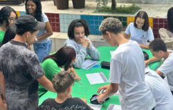 #paratodosverem: Em um pátio externo, cerca de 12 jovens (estudantes) estão reunidos em torno de uma mesa de cimento pintada de verde. Eles estão sentados e em pé, escrevendo em folhas de papel com grades. Alguns vestem camisetas de uniformes verdes e brancas. Há estojos e canetas sobre a mesa. Ao fundo, veem-se bancos com azulejos coloridos (vermelho, azul, verde, amarelo) e vasos de plantas.