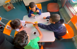#paratodosverem: Quatro crianças sentadas ao redor de uma mesa branca quadrada realizam uma atividade de plantio. Cada uma tem um copinho de plástico com terra. No centro da mesa, há um recipiente amarelo onde algumas crianças colocam as mãos para pegar sementes. As cadeiras da sala são amarelas e alaranjadas, e o chão é azul. Na parede ao fundo, há pilhas de livros sobre uma mesa.