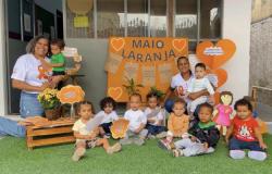 #paratodosverem: Crianças e educadoras participam de atividade do Maio Laranja em espaço escolar decorado com cartazes e corações na cor laranja. Ao fundo há mensagens de conscientização sobre proteção da infância, enquanto os participantes posam sentados sobre gramado sintético. 