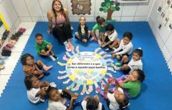 #paratodosverem: Crianças e uma professora sentadas em círculo sobre um tapete azul participam de atividade em sala. No centro, há um cartaz sobre o Dia da Síndrome de Down, com desenhos de meias coloridas e de corações nas cores azul e amarela, além da frase: “Ser diferente é o que torna o mundo mais bonito”.