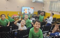 #paratodosverem: Em uma sala com cadeiras pretas organizadas em fileiras, várias crianças usam uniformes escolares verdes e brancos e sorriem para a câmera. Outras crianças também uniformizadas seguram pequenas plaquinhas com frases de cuidado aos livros. À direita, uma professora de vestido preto e óculos sorri sentada em uma das cadeiras. Ao fundo, há uma televisor com uma imagem projetada