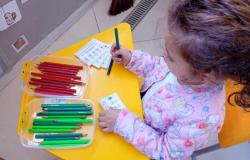#paratodosverem: Em uma mesa amarela, uma criança segura um lápis enquanto avalia a refeição oferecida pela creche. Sobre a mesa, há cartões ilustrados e recipientes com lápis das cores vermelha e verde.