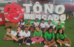 #paratodosverem: Grupo de crianças posa sentado à frente do letreiro “TÔ NO MERCADO”, em um espaço com estrutura metálica e piso verde. Os alunos usam uniforme escolar com blusas nas cores branca e verde e sorriem para a foto durante a visita ao Mercado.