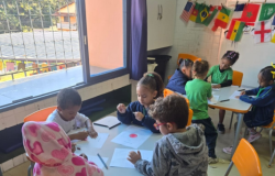 #paratodosverem: Crianças pequenas sentadas em mesas brancas com cadeiras laranja desenham a bandeira do Japão em folhas de papel numa sala de aula. À esquerda, uma janela com grade de ferro preta e cortina roxa. Na parede do fundo, um varal com bandeiras de vários países, incluindo Brasil, EUA, Camarões e Senegal.
