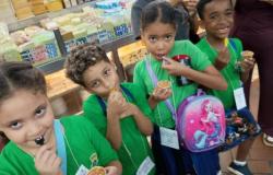 #paratodosverem: Quatro crianças, dois meninos e duas crianças, usando uniformes escolares da Prefeitura de Belo Horizonte e crachás, provam doce no Mercado Central. Cada uma segura um pequeno copo com doce de leite e colher, enquanto ao fundo aparecem balcões refrigerados com queijos, etiquetas de preços e produtos expostos