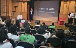 #paratodosverem: a imagem mostra um auditório com vários(as) estudantes sentados(as) em cadeiras estofadas, assistindo a uma palestra.