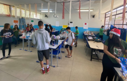 #paratodosverem: a foto mostra uma sala com paredes pintadas em azul claro e branco, decorada com tiras de papel penduradas no teto. Há várias mesas organizadas em fileiras, sobre as quais estão expostos diversos produtos, como bolas de futebol, fones de ouvido, doces, calçados, itens de maquiagem e kits embrulhados em celofane. Vários(as) estudantes circulam pela sala, observando os itens. 