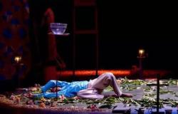 Cena do espetáculo teatral "Capelas Gerais". Um ator está deitado de costas no chão do palco, cercado por folhas verdes e flores espalhadas. Ele veste blusa azul de mangas compridas e calça clara. Acima dele, há uma tigela de vidro com água equilibrada sobre um suporte alto. Ao fundo, o cenário escuro traz velas acesas e uma cruz no canto inferior direito.