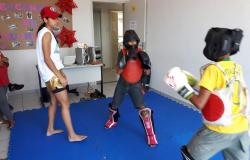 Dois alunos devidamente equipados praticam Muay Thai sob supervisão da professora, em um tatami.