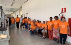 Dezessete idosos de camiseta laranja com os dizeres: "Alegria de viver" visitam Museu de Artes e Ofícios. 
