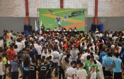Mais de cem estudantes em frente a mesa com seis pessoas sentadas à mesa, uma no púlpito à esquerda e, ao fundo, banner dos Jogos Escolares de Belo Horizonte. 