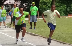 Dois atletas correm e três pessoas observam, durante o dia. 