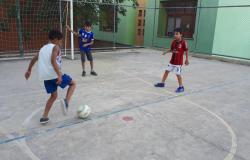 Três garotos jogam futebol em quadra, durante o dia. 