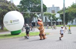 Homem fantasiado de dragão caminha em rua fechada, ao lado de bola com a logo da Prefeitura; pessoas caminham ao redor, durante o dia. 