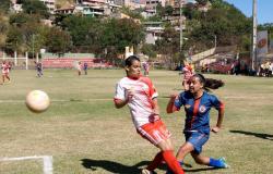 Duas garotas de times diferentes jogam futebol em campo; à direita, a bola está no ar. 