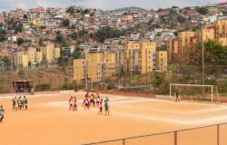 Times jogam em quadra de futebol de sem grama com cidade ao fundo. 